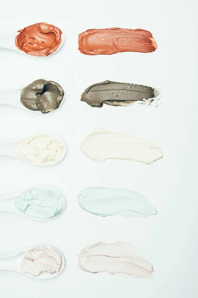Top view of spoons and various colorful clay mask smudges placed in row isolated on white surface — Stock Photo