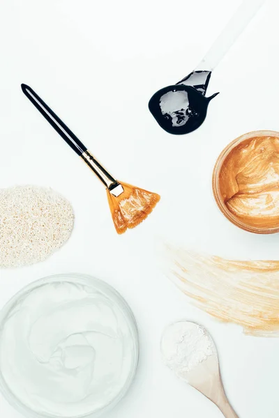 Top view of sponge, spoon with clay powder, containers with clay masks, smudge and spoon with black and golden mask isolated on white surface — Stock Photo