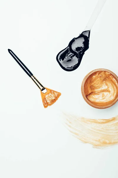 Vista superior de máscara dourada em recipiente, escova, mancha e colher com máscara preta isolada na superfície branca — Fotografia de Stock