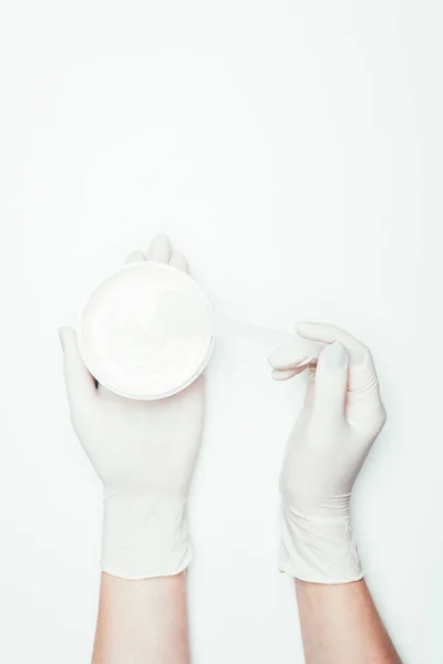 Image recadrée de la femme dans des gants en latex tenant récipient avec masque en argile et cuillère isolée sur blanc — Photo de stock