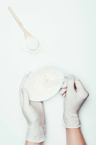 Cropped shot of woman in latex gloves with plate with clay mask on surface with spoon and clay powder — Stock Photo