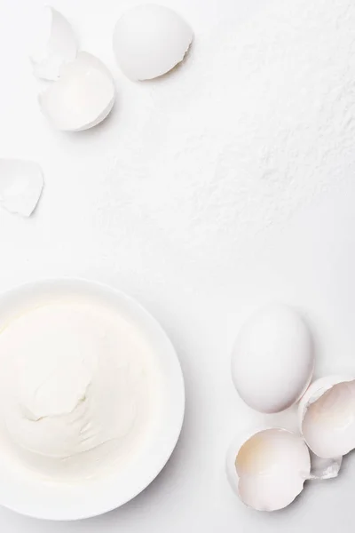 Vista superior de creme azedo na tigela e cascas de ovo rachado na superfície branca derramada com farinha — Fotografia de Stock