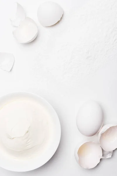 Top view of sour cream in bowl and cracked egg shells on white surface spilled with flour — Stock Photo