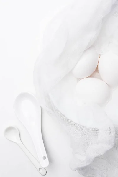 Top view of raw chicken eggs in cheesecloth with spoons on white surface — Stock Photo