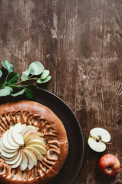 Blick von oben auf hausgemachten Apfelkuchen und frische Äpfel mit grünen Blättern auf Holztischplatte — Stockfoto