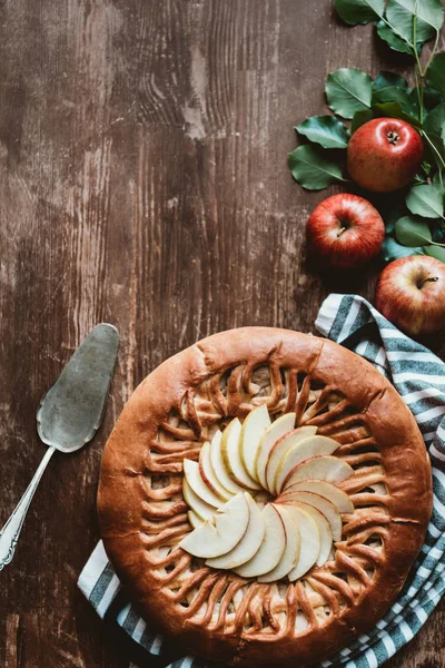 Vista superior da torta de maçã arranjada, servidor de bolo e maçãs frescas com folhas verdes na superfície de madeira — Fotografia de Stock