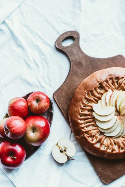 Draufsicht auf frische Äpfel und hausgemachten Apfelkuchen auf Holzschneidebrett auf weißer Tischdecke — Stockfoto