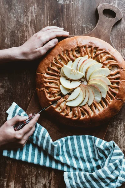 Partial view of woman cutting homemade apple pie on cutting board on wooden tabletop — Stock Photo