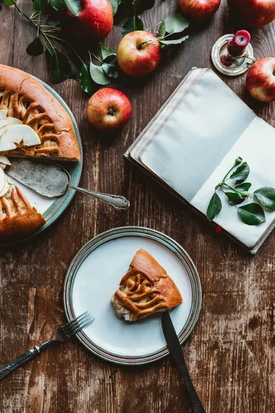 Flat lay with piece of homemade apple pie, cutlery, empty notebook and fresh apples arranged on wooden surface — Stock Photo