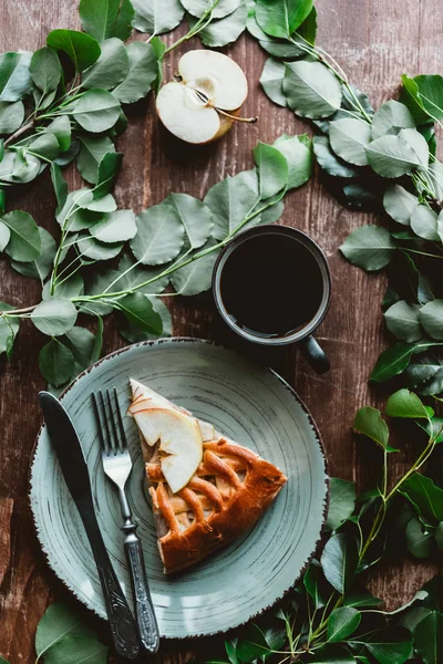 Top view of piece of apple pie, cup of coffee and green leaves arranged on wooden surface — Stock Photo
