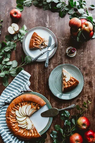 Flat lay with pieces of apple pie on plates, cutlery, linen, green leaves and fresh apples arranged on wooden tabletop — Stock Photo