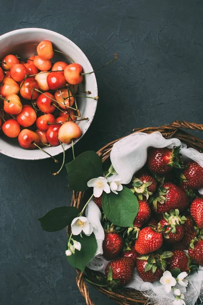 Top view of sweet cherries in bowl and ripe strawberries with jasmine flowers in basket on black — Stock Photo