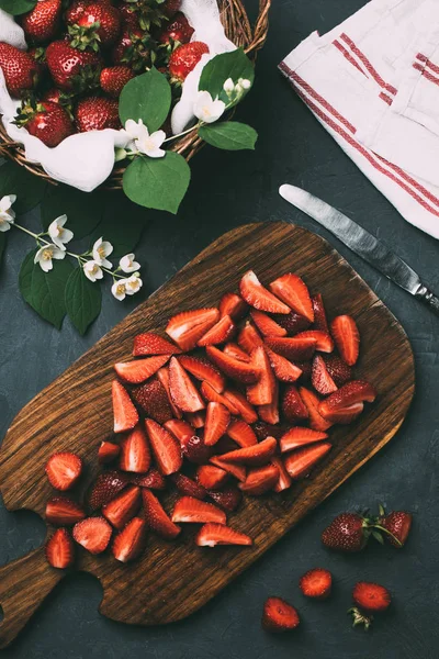 Vue de dessus des fraises tranchées sur planche à découper en bois et des fleurs de jasmin sur fond noir — Photo de stock
