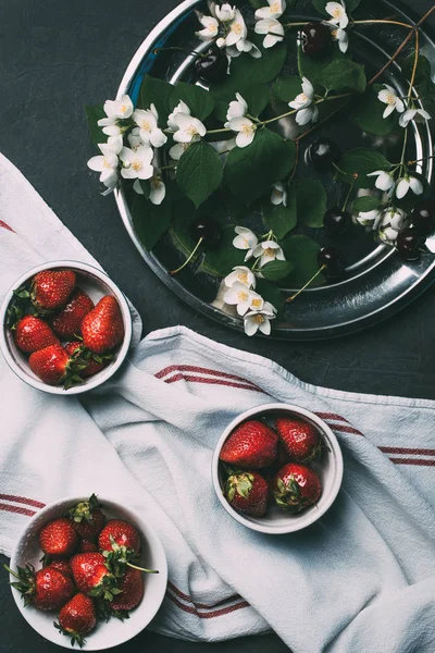 Vue de dessus des fraises rouges mûres dans des bols, serviette et fleurs de jasmin sur noir — Photo de stock