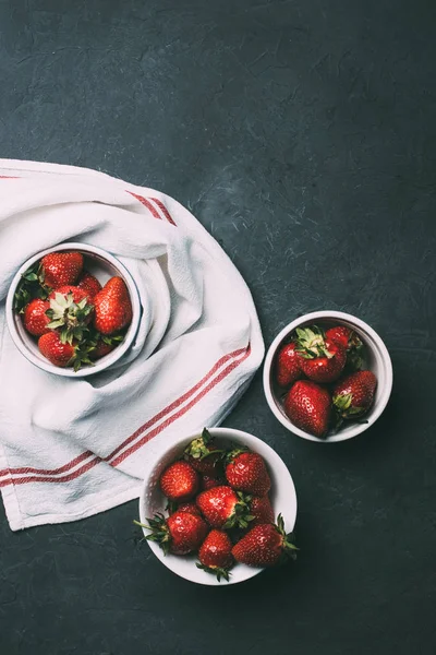 Vue de dessus des fraises rouges mûres dans des bols et serviette sur noir — Photo de stock