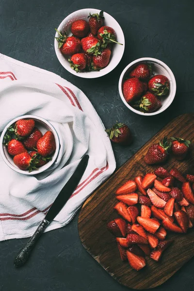 Top view or ripe whole strawberries in bowls and sliced strawberries on wooden cutting board on black — Stock Photo