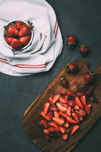Top view or ripe whole strawberries in bowl and sliced strawberries on wooden cutting board on black — Stock Photo