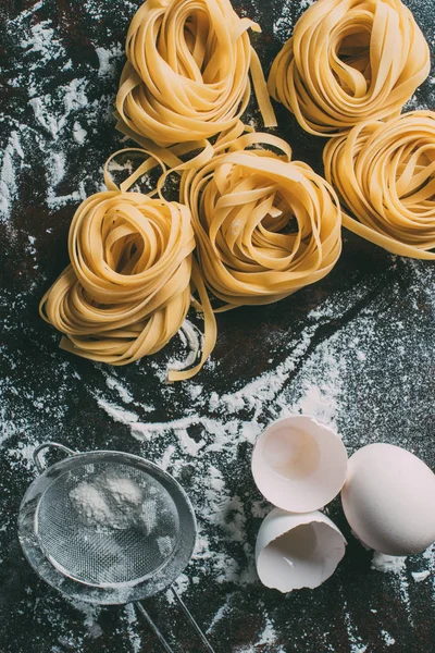 Draufsicht auf rohe Tagliatelle-Pasta, Sieb, Ei und Eierschale auf Tisch mit Mehl bedeckt — Stockfoto