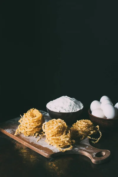 Vista frontal de la tabla de cortar con pasta cruda, cuencos con harina y huevos en la mesa sobre fondo negro - foto de stock