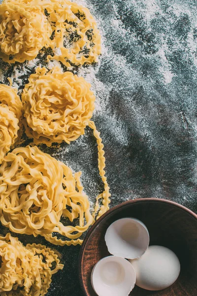 Elevated view of raw pasta, bowl with egg and egg shell on table covered by flour — Stock Photo