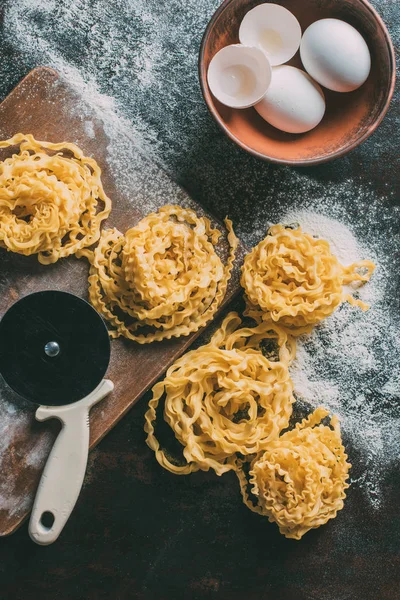 Top view of adjustable dough cutter, raw pasta, cutting board, bowl with egg shell and eggs on table covered by flour — Stock Photo