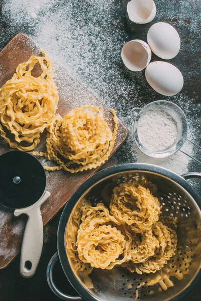 Vue surélevée de coupe-pâte réglable, pâtes crues, passoire, tamis, coquilles d'œufs et oeufs sur la table couverte de farine — Photo de stock