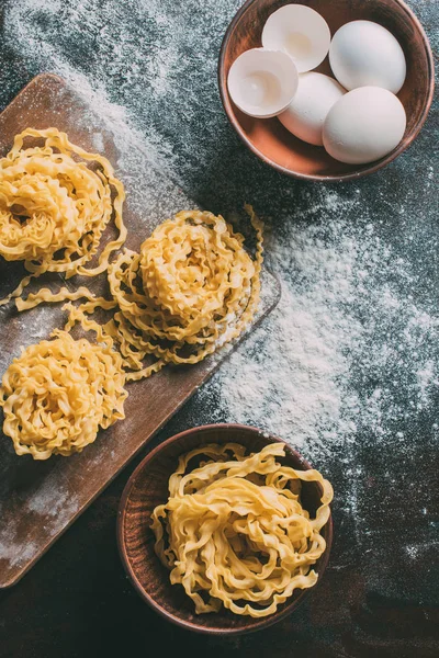 Vue du dessus de la planche à découper, des bols, des œufs, des coquilles d'œufs et des pâtes crues sur la table couverte de farine — Photo de stock