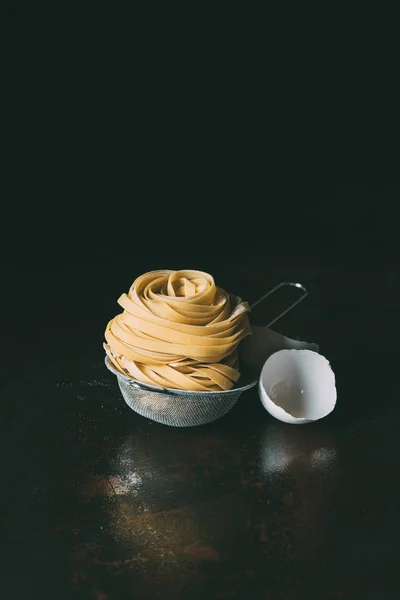 Close up view of tagliatelle pasta in sieve and egg shell on black background — Stock Photo