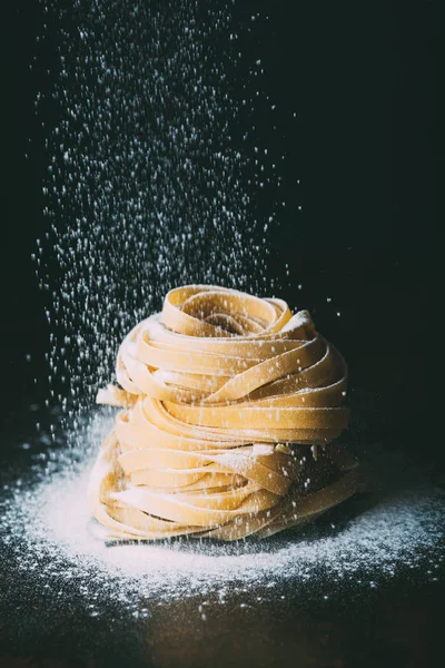 Nahaufnahme von Mehl, das auf rohe Tagliatelle-Pasta auf schwarzem Hintergrund fällt — Stockfoto