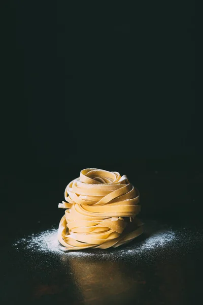 Nahaufnahme von rohen Tagliatelle-Pasta und Mehl auf dem Tisch auf schwarzem Hintergrund — Stockfoto