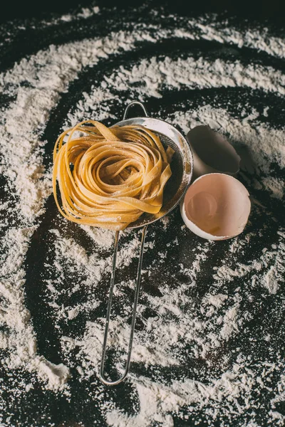 Close up image of raw tagliatelle pasta, sieve and egg shells on table covered by flour — Stock Photo