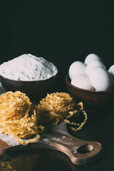 Cerrar imagen de pasta cruda, cuencos, harina, huevos y tabla de cortar sobre fondo negro - foto de stock