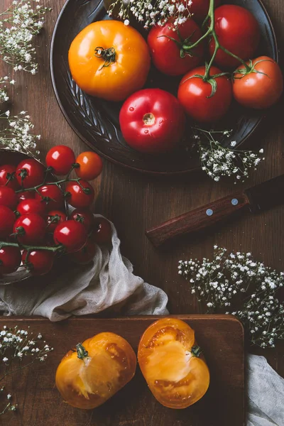 Vista superior de diferentes tomates na placa de cerâmica e placa de corte com faca no fundo de madeira — Fotografia de Stock