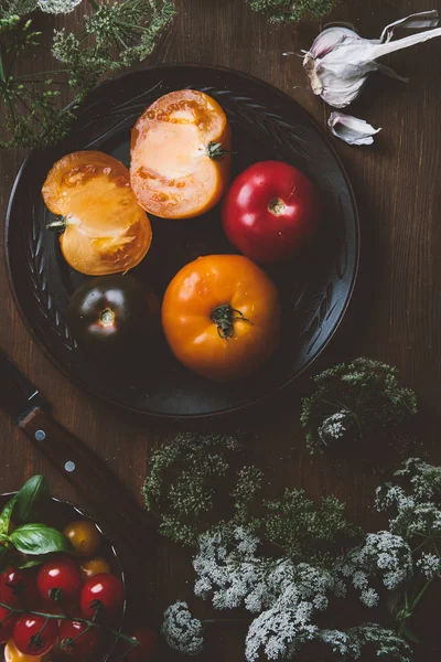 Vista superior de tomates en placa de cerámica con cuchillo sobre fondo de madera con flores de perejil - foto de stock