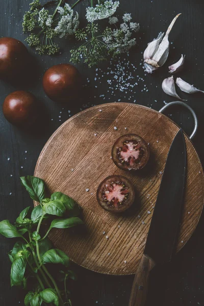 Top view of black prince tomatoes on cutting board with knife, garlic, salt, basil leaves and flowers — Stock Photo