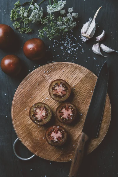 Top view of black prince tomatoes on cutting board with knife, garlic, salt and flowers — Stock Photo