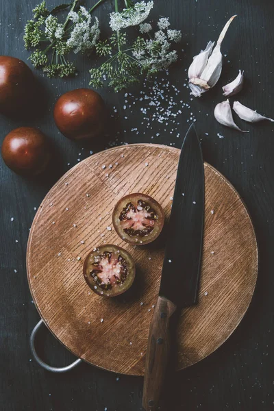 Top view of black prince tomatoes on wooden cutting board with knife, garlic, salt and flowers — Stock Photo
