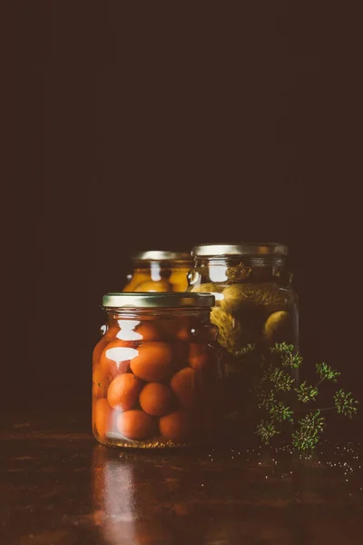 Glass jars with preserved tomatoes, cucumbers and dill on wooden table in dark kitchen — Stock Photo