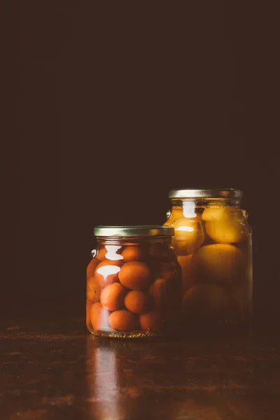 Glass jars with preserved red and yellow tomatoes on wooden table in dark kitchen — Stock Photo