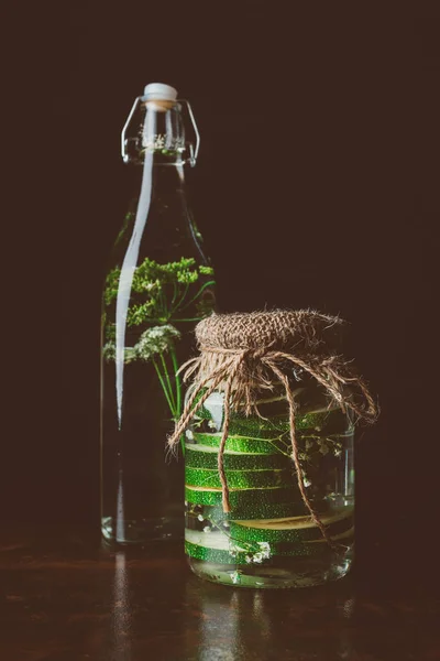 Bouteille en verre avec eau et aneth et bocal en verre avec courgettes sur table en bois sur noir — Photo de stock