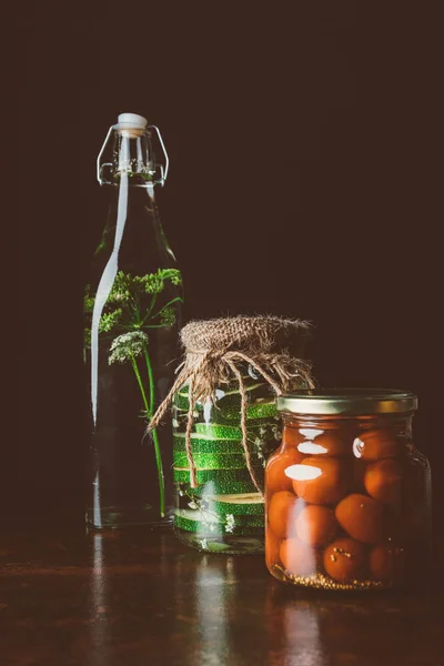 Jarros de vidro com tomates preservados e abobrinha em mesa de madeira na cozinha escura — Fotografia de Stock