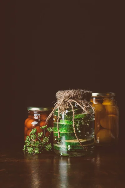 Glass jars with preserved vegetables on wooden table in dark kitchen — Stock Photo