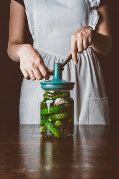 Imagen recortada de la mujer preparando pepinos preservados en frasco de vidrio en la mesa de madera - foto de stock