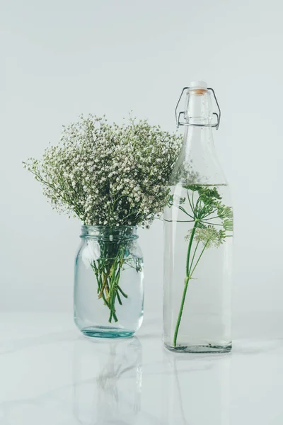 Glass bottle with water and dill and glass jar with flowers on white table — Stock Photo