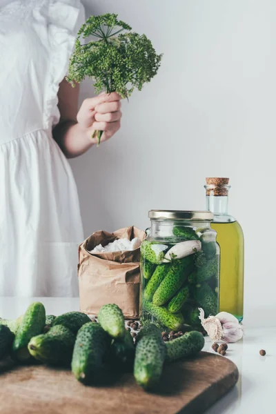 Cropped image of woman preparing preserved organic cucumbers and holding dill at kitchen — Stock Photo