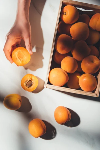 Partial view of woman holding piece of apricot with wooden box full of apricots near by on light marble surface — Stock Photo