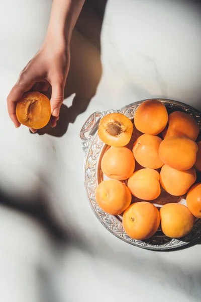 Partial view of woman holding piece of apricot with metal tray full of ripe apricots near by on light marble surface — Stock Photo
