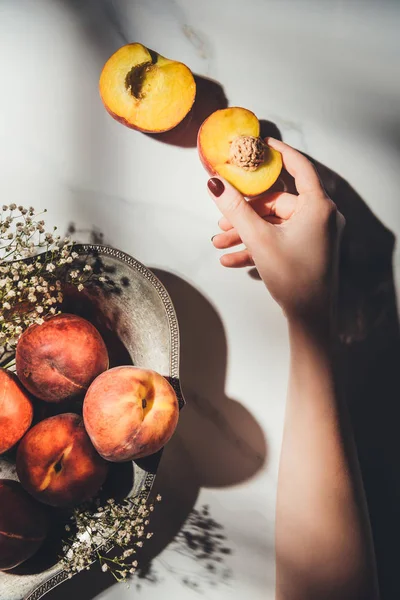 Tiro cortado de mulher com pedaço de pêssego na mão e tigela cheia de pêssegos maduros e flores de gypsophila perto de em mesa de mármore leve — Fotografia de Stock