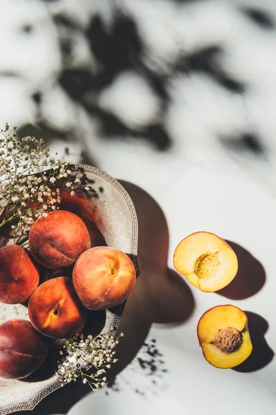 Flat lay com pêssegos maduros em tigela de metal com flores de gypsophila na superfície de mármore leve com sombras — Fotografia de Stock