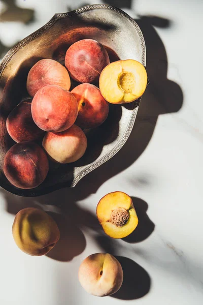 Flat lay with ripe peaches in metal bowl on light marble surface with shadows — Stock Photo
